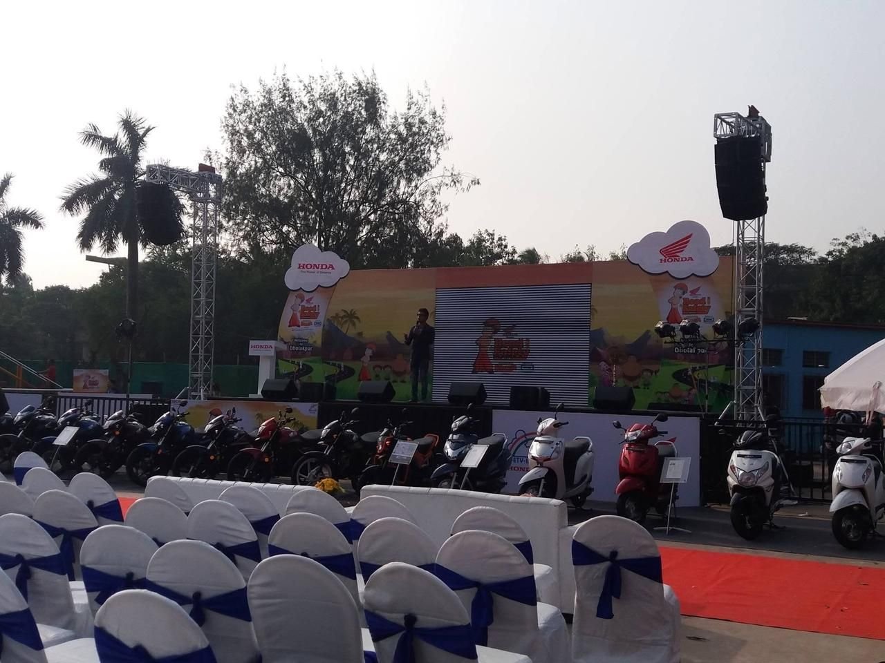 Outdoor dealer meet with covered seating, illustrated stage backdrop and a two-wheeler product line-up