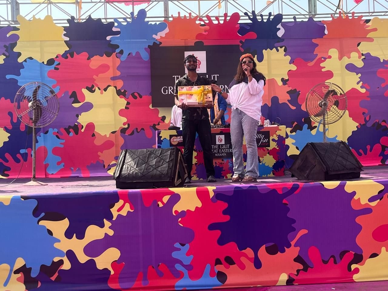 Hosts on a festival stage with a colour-splash backdrop and stage monitors